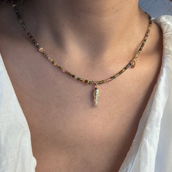 A close-up of a person's neck and chest wearing a delicate beaded necklace with a small gold feather pendant. The necklace features small multicolored beads and gold-toned metal details, worn over a white top.