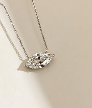 A silver chain necklace with a marquise-cut clear gemstone pendant, displayed on a warm beige background with soft shadows.