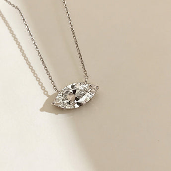 A silver chain necklace with a marquise-cut clear gemstone pendant, displayed on a warm beige background with soft shadows.