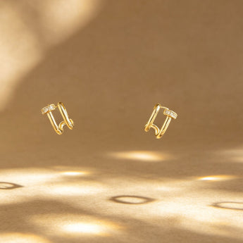 A pair of small gold hoop-style earrings with tiny clear stones, shown floating against a warm beige background with soft shadow patterns.