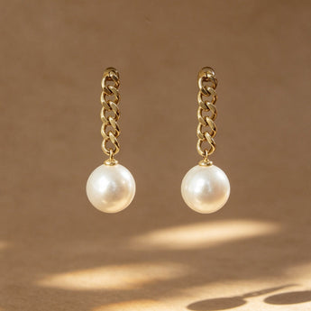 A pair of gold chain drop earrings with white pearl pendants, displayed against a warm brown background with artistic leaf shadows.