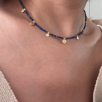 A close-up of a person's neck and upper chest wearing a delicate necklace made of dark blue beads with small gold coin charms and a central gold disc pendant.