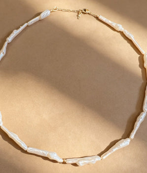 A necklace made of irregular white shell-like beads with a gold clasp, arranged in a circle on a warm beige background with soft diagonal shadows.