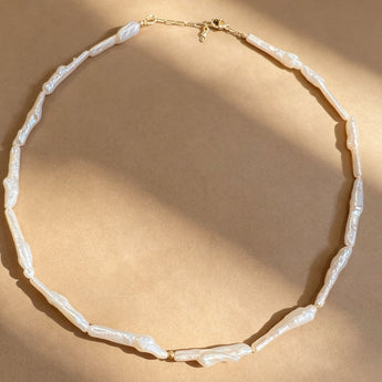 A necklace made of irregular white shell-like beads with a gold clasp, arranged in a circle on a warm beige background with soft diagonal shadows.