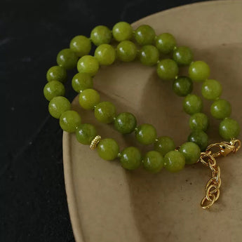 A beaded strand bracelet made of round green beads with a small gold spacer and a gold chain clasp, resting on a beige ceramic dish against a dark background.