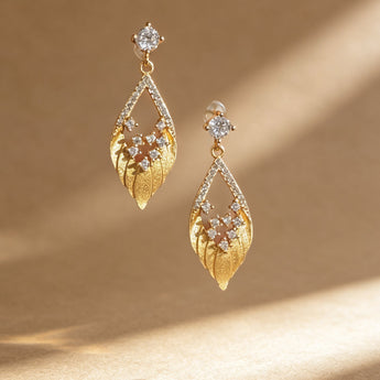A pair of gold dangle earrings with leaf-like textured drops, clear gemstone accents, and a small clear round stone at the top, displayed on a warm beige background with soft diagonal shadows.