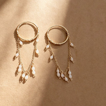 A pair of gold-toned dangle earrings with circular hoop tops and multiple dangling chain sections adorned with small white pearl-like beads, photographed on a warm beige textured surface with diagonal light shadows.
