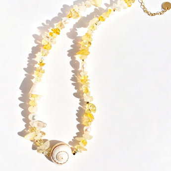 A delicate necklace made of translucent yellow and white flower-shaped beads, a spiral shell pendant, and a gold-tone clasp, displayed on a clean white background with soft shadows.
