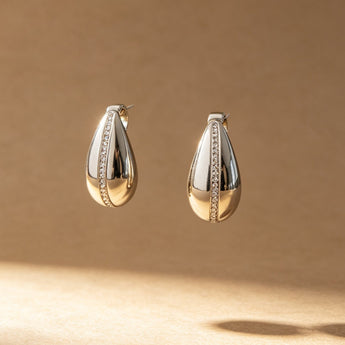 A pair of polished silver teardrop hoop earrings with a central channel of small clear stones, displayed against a warm brown background with dramatic diagonal shadows.