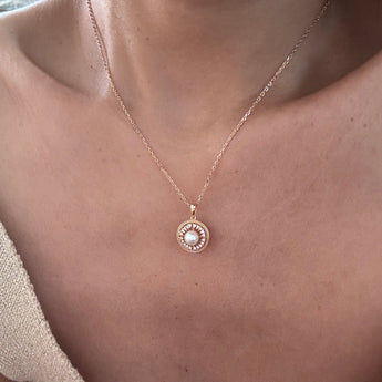 A close-up of a person's chest and neck wearing a delicate rose gold chain necklace with a circular pendant featuring a white pearl surrounded by small clear stones.