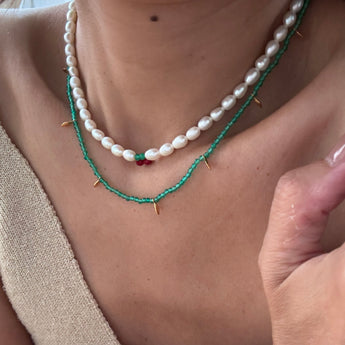 Close-up of a person wearing a layered necklace set: one strand of irregular white pearls and one turquoise beaded chain with small gold spikes and a tiny red bead accent.