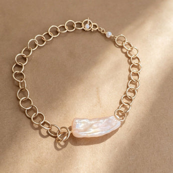 A delicate gold chain bracelet with a large, irregular pearlescent white shell centerpiece, displayed on a textured brown background with soft diagonal shadows.