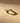 A textured gold band ring with a unique patterned design, resting on a warm beige surface with dramatic lighting and a soft shadow.