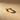 A textured gold band ring with a unique patterned design, resting on a warm beige surface with dramatic lighting and a soft shadow.