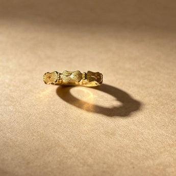 A textured gold band ring with a unique patterned design, resting on a warm beige surface with dramatic lighting and a soft shadow.