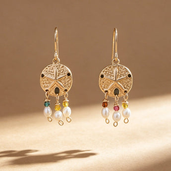A pair of gold-toned dangling earrings with circular filigree tops and small multicolored bead accents above white pearl drops, photographed on a warm beige surface with soft shadows.