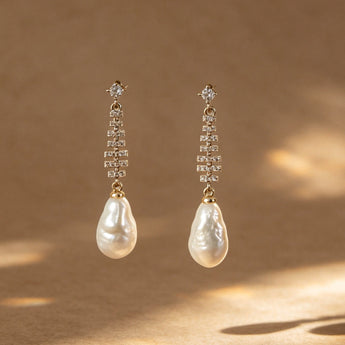 A pair of gold drop earrings with baroque white freshwater pearls and small clear stones, displayed against a warm brown textured background with artistic leaf shadows.