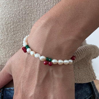 A close-up of a person's wrist wearing a beaded stretch bracelet with white baroque pearls and small red and green beads, styled with a beige knit sweater and blue jeans.