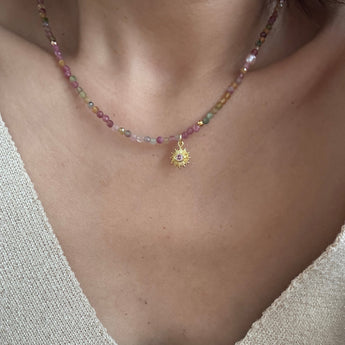 A close-up of a person wearing a delicate beaded necklace with a small gold sunburst pendant. The necklace features small, faceted beads in pink, green, and clear tones, worn over a beige knit V-neck top.
