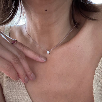 A close-up of a person wearing a delicate silver chain necklace with a small square-cut clear gemstone pendant, paired with a beige knit top.