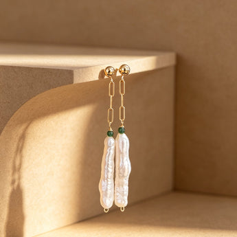 A pair of long dangling earrings with gold-toned chain links, small green beads, and irregular white pearl-like drops, hanging against a warm beige studio set with strong sunlight and shadows.