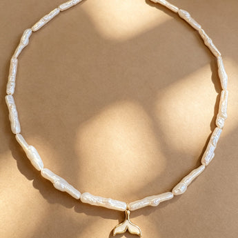 A gold chain necklace featuring irregular white faux-pearl beads and a gold-plated whale tail pendant, displayed on a warm beige background with soft shadow overlays.