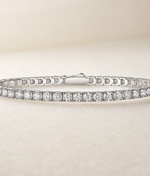 A silver tennis bracelet with a continuous row of clear round-cut gemstones, displayed on a soft beige textured surface.