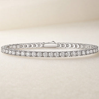 A silver tennis bracelet with a continuous row of clear round-cut gemstones, displayed on a soft beige textured surface.