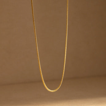 A delicate gold chain necklace hanging in a U-shape against a warm beige background, with soft lighting casting a shadow on the surface below.