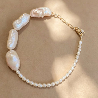 A baroque pearl necklace with a gold chain extender and clasp, displayed on a textured beige background with soft shadows.