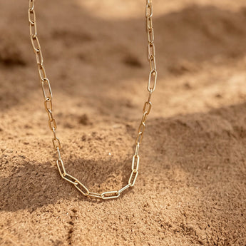 A gold paperclip chain necklace laid on sandy ground in warm natural light, with a blurred earthy background and shadows.