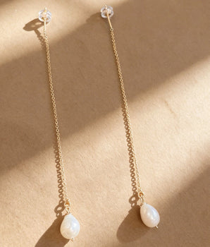 A pair of long gold dangle earrings with clear crystal studs and teardrop-shaped white pearl-like drops, displayed on a textured beige surface with diagonal shadows.
