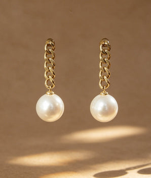 A pair of gold chain drop earrings with white pearl pendants, displayed against a warm brown background with artistic leaf shadows.