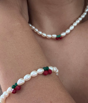 A close-up of a person's neck and arm wearing a matching jewelry set: a pearl necklace and a pearl bracelet, both decorated with small red berry charms and green leaf accents.