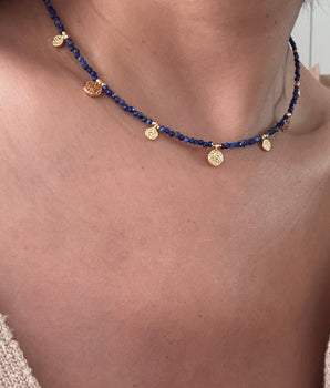 A close-up of a person's neck and upper chest wearing a delicate necklace made of dark blue beads with small gold coin charms and a central gold disc pendant.