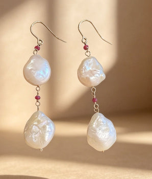 A pair of dangle earrings with irregular white baroque pearls, small red gemstone accents, and silver hooks, displayed against a warm beige background with dramatic window light shadows.