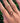 A close-up of a hand wearing three delicate gold rings outdoors against a background of green foliage. The rings include a leaf motif, a small floral cluster with red and green stones, and a thin gold band.