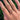 A close-up of a hand wearing three delicate gold rings outdoors against a background of green foliage. The rings include a leaf motif, a small floral cluster with red and green stones, and a thin gold band.