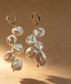 A pair of gold hoop earrings with irregular white baroque pearls, shown floating against a warm brown background with dramatic shadows.