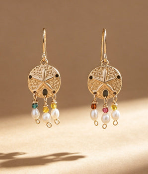 A pair of gold-toned dangling earrings with circular filigree tops and small multicolored bead accents above white pearl drops, photographed on a warm beige surface with soft shadows.