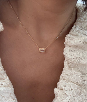 A close-up of a person wearing a delicate gold chain necklace with a small rectangular pendant featuring a clear gemstone, styled with a white lace top.