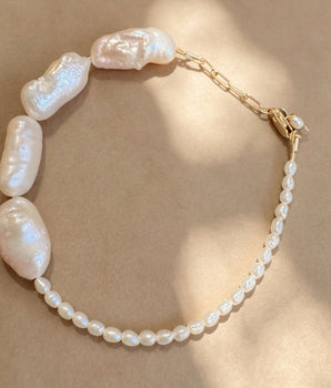 A baroque pearl necklace with a gold chain extender and clasp, displayed on a textured beige background with soft shadows.